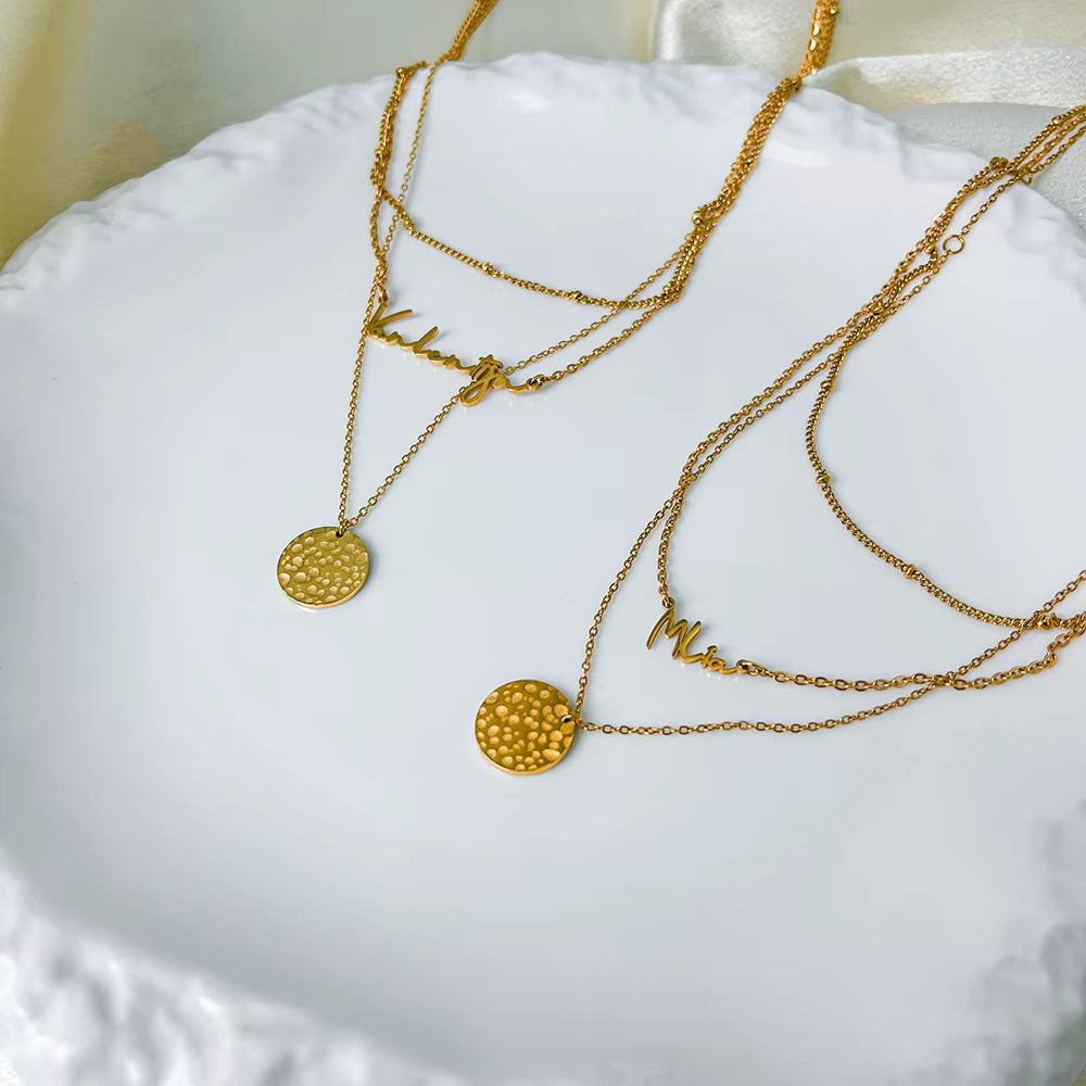 Gold stainless steel multilayer necklace with custom name pendant, bead chain, and textured disc charm displayed on a white surface.