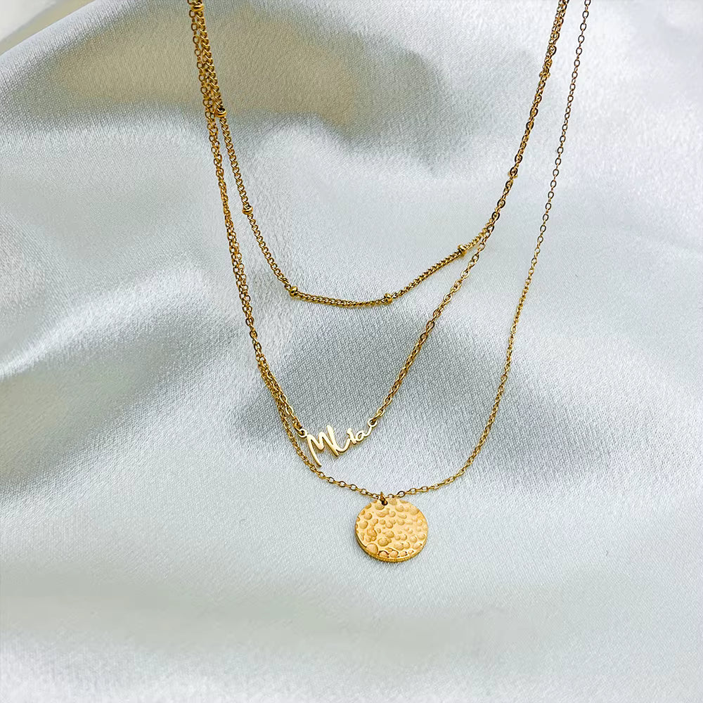 Gold stainless steel multilayer necklace with custom name pendant, bead chain, and textured disc charm displayed on a white surface.