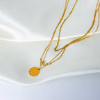 Gold stainless steel multilayer necklace with custom name pendant, bead chain, and textured disc charm displayed on a white surface.