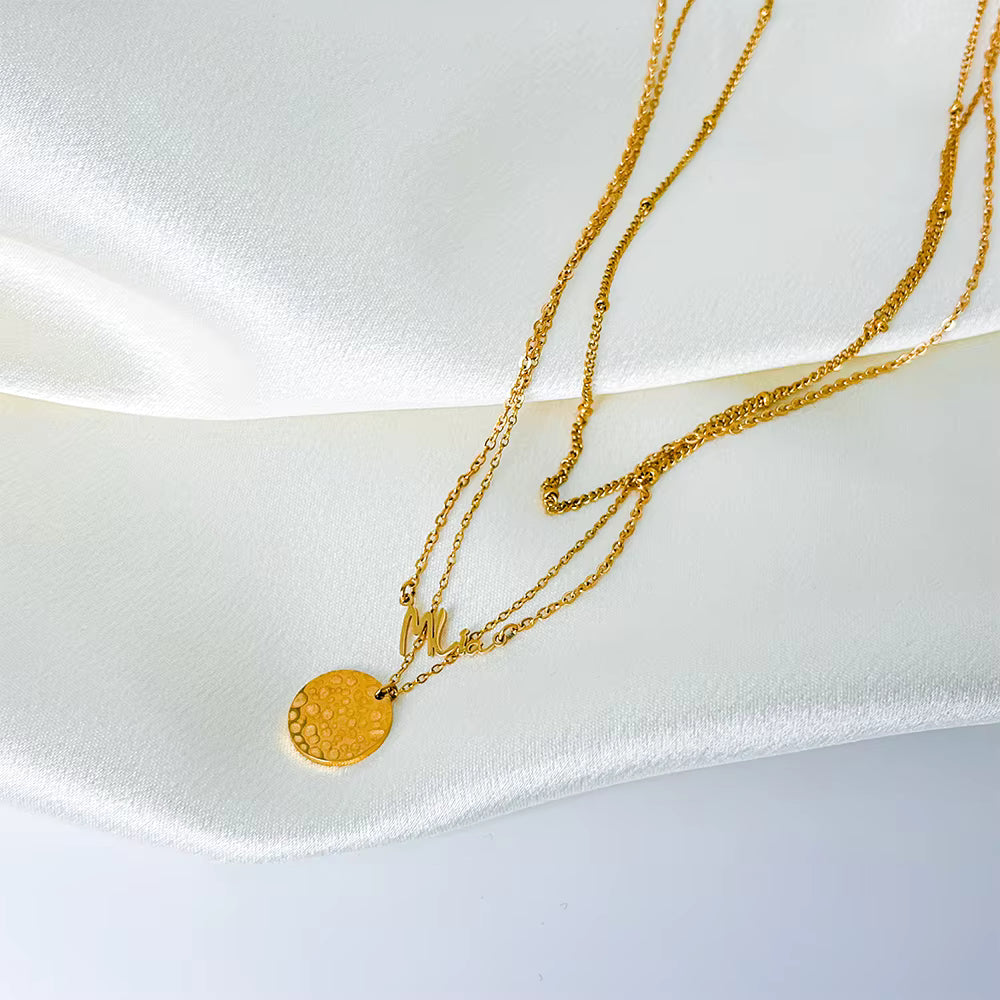 Gold stainless steel multilayer necklace with custom name pendant, bead chain, and textured disc charm displayed on a white surface.