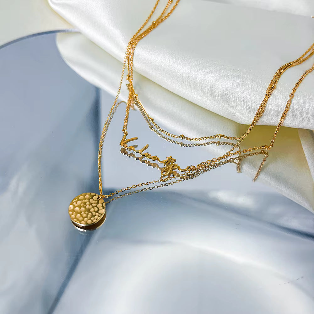 Gold stainless steel multilayer necklace with custom name pendant, bead chain, and textured disc charm displayed on a white surface.