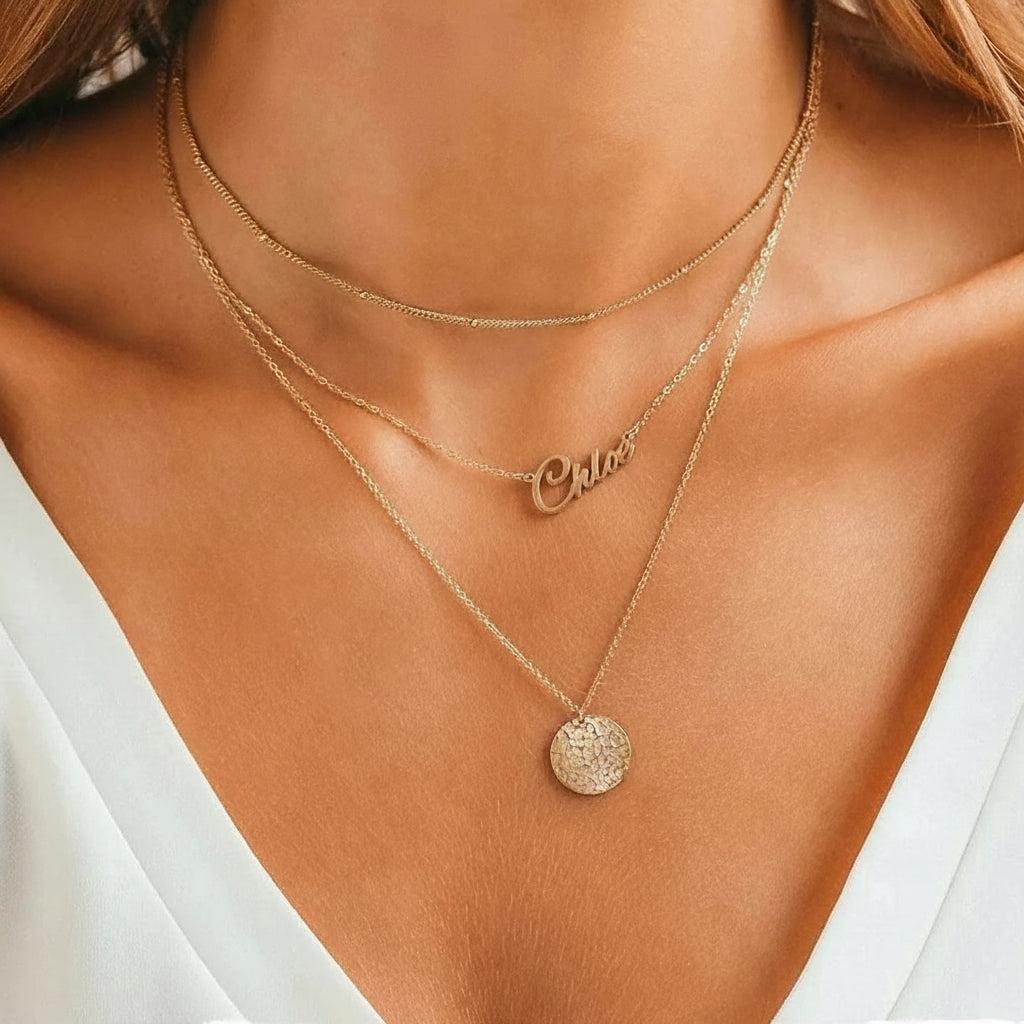 Gold stainless steel multilayer necklace with custom name pendant, bead chain, and textured disc charm displayed on a white surface.