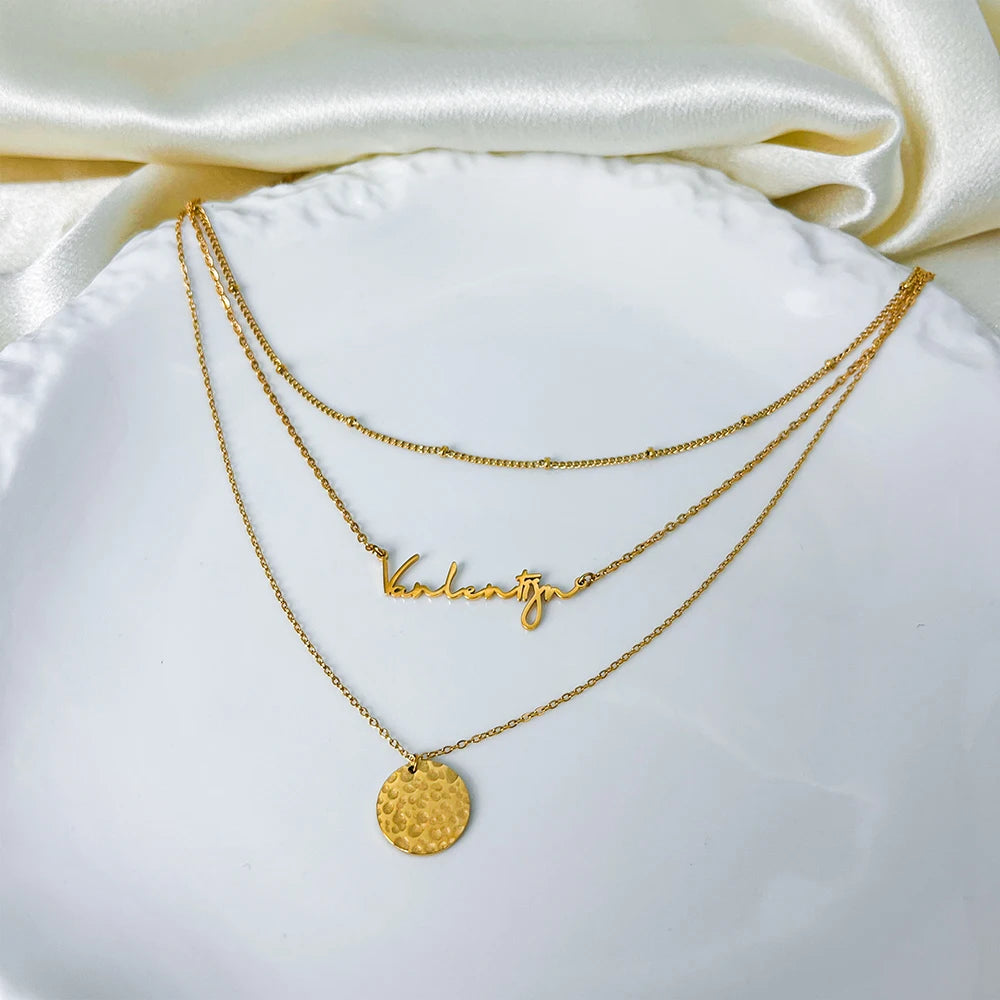 Gold stainless steel multilayer necklace with custom name pendant, bead chain, and textured disc charm displayed on a white surface.